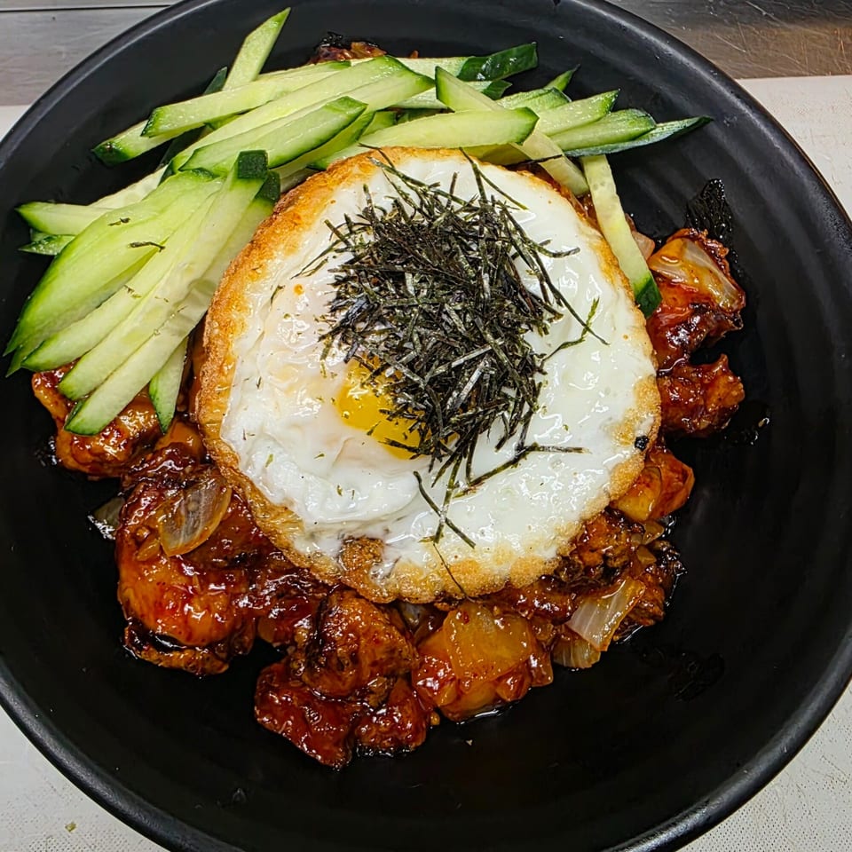 Japanese dish with glazed chicken, a fried egg topped with seaweed, and cucumber slices in a black bowl.