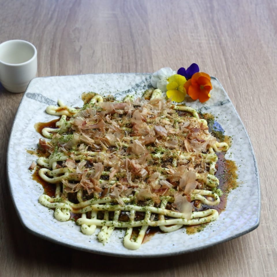 Okonomiyaki topped with mayonnaise, brown sauce, bonito flakes, and edible flowers on a white plate.