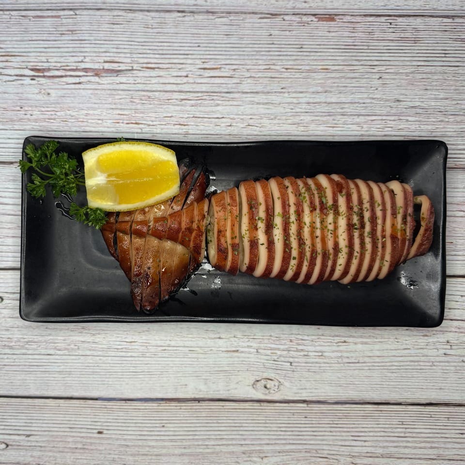 Grilled whole squid, sliced and garnished with parsley and a lemon wedge on a black plate.