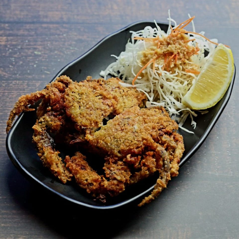 Crispy fried soft-shell crab, shredded cabbage salad, and a lemon wedge on a black plate.